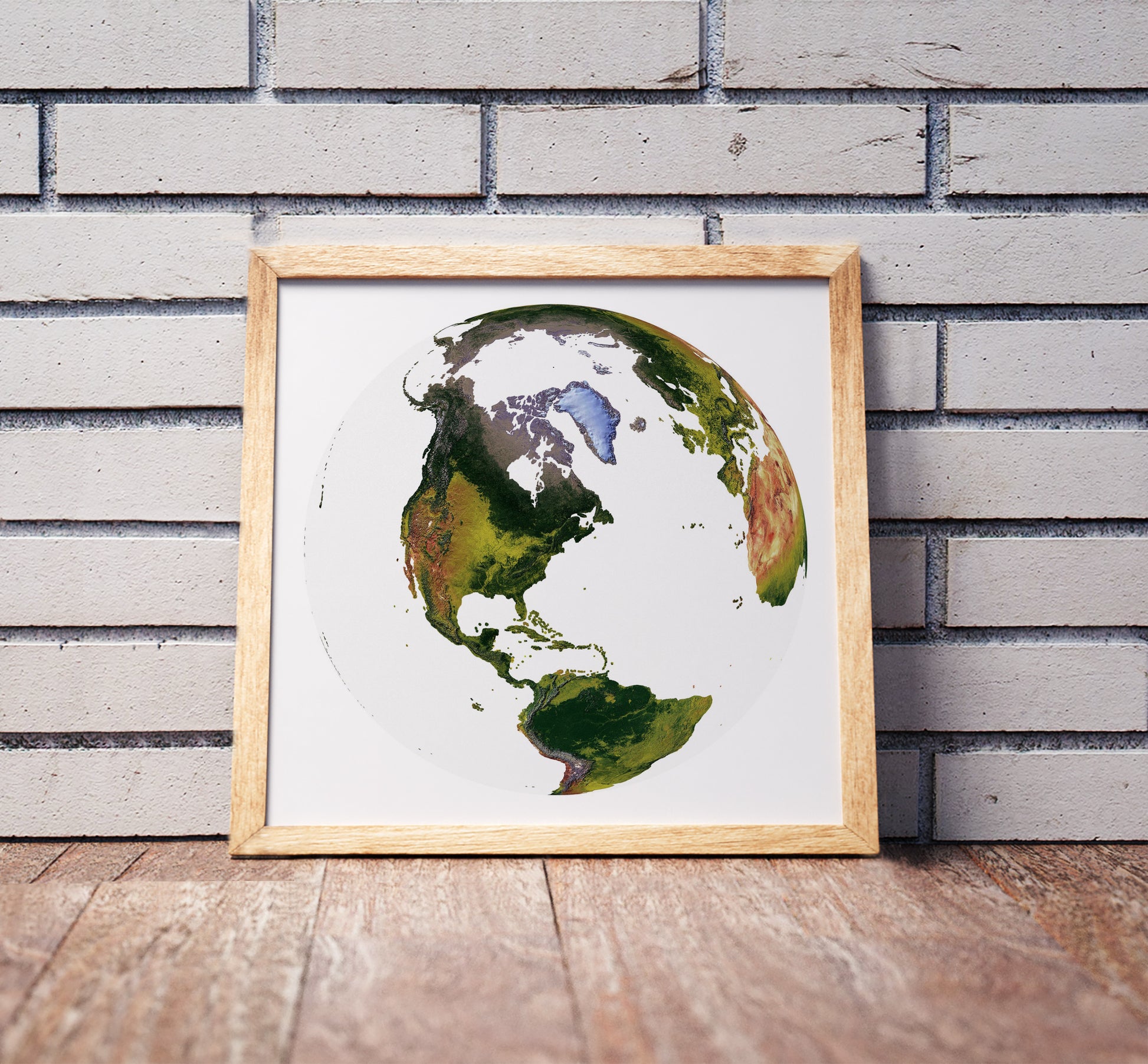 The World From Space projection shaded relief world map in a wooden frame. Realistic terrain art print for home decor, displayed against a brick wall