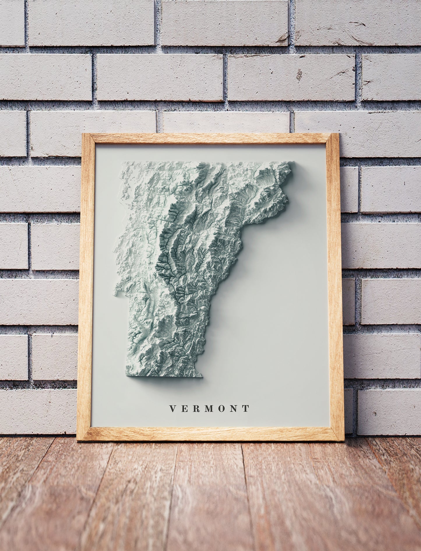 Vermont shaded relief map in a wooden frame. Realistic terrain art print for home decor, displayed against a brick wall