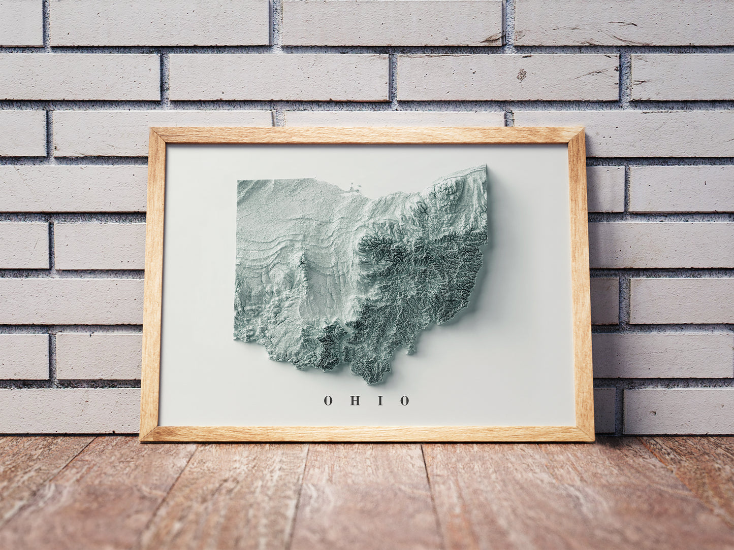 Ohio shaded relief map in a wooden frame. Realistic terrain art print for home decor, displayed against a brick wall