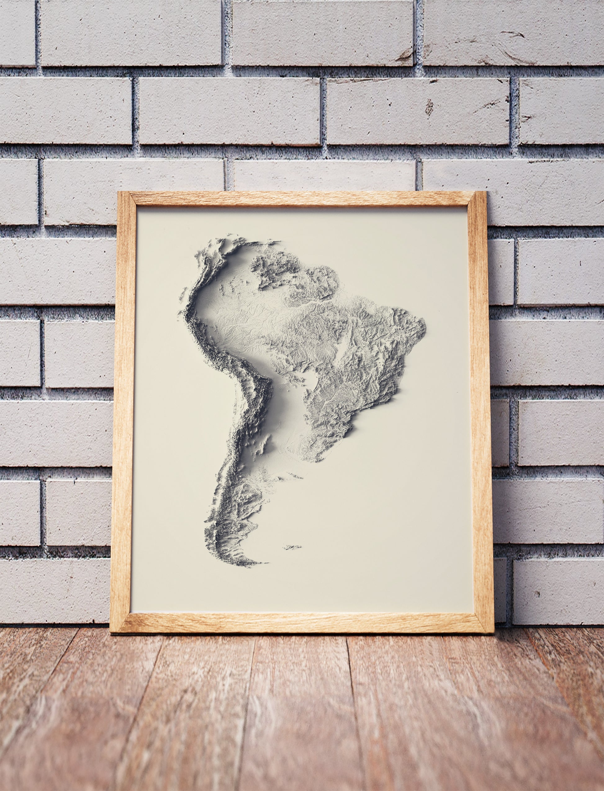 South America shaded relief map in a wooden frame. Realistic terrain art print for home decor, displayed against a brick wall