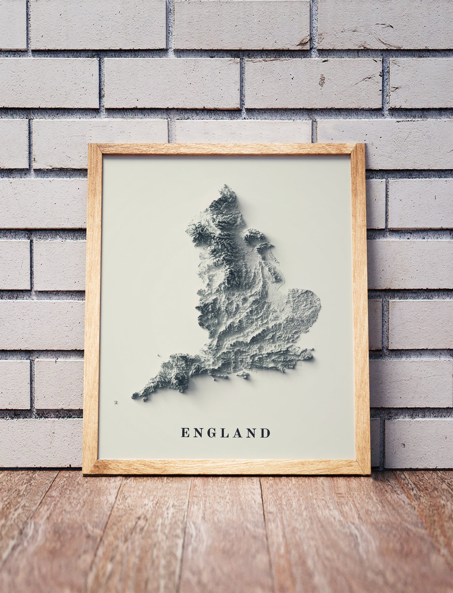 England shaded relief map in a wooden frame. Realistic terrain art print for home decor, displayed against a brick wall