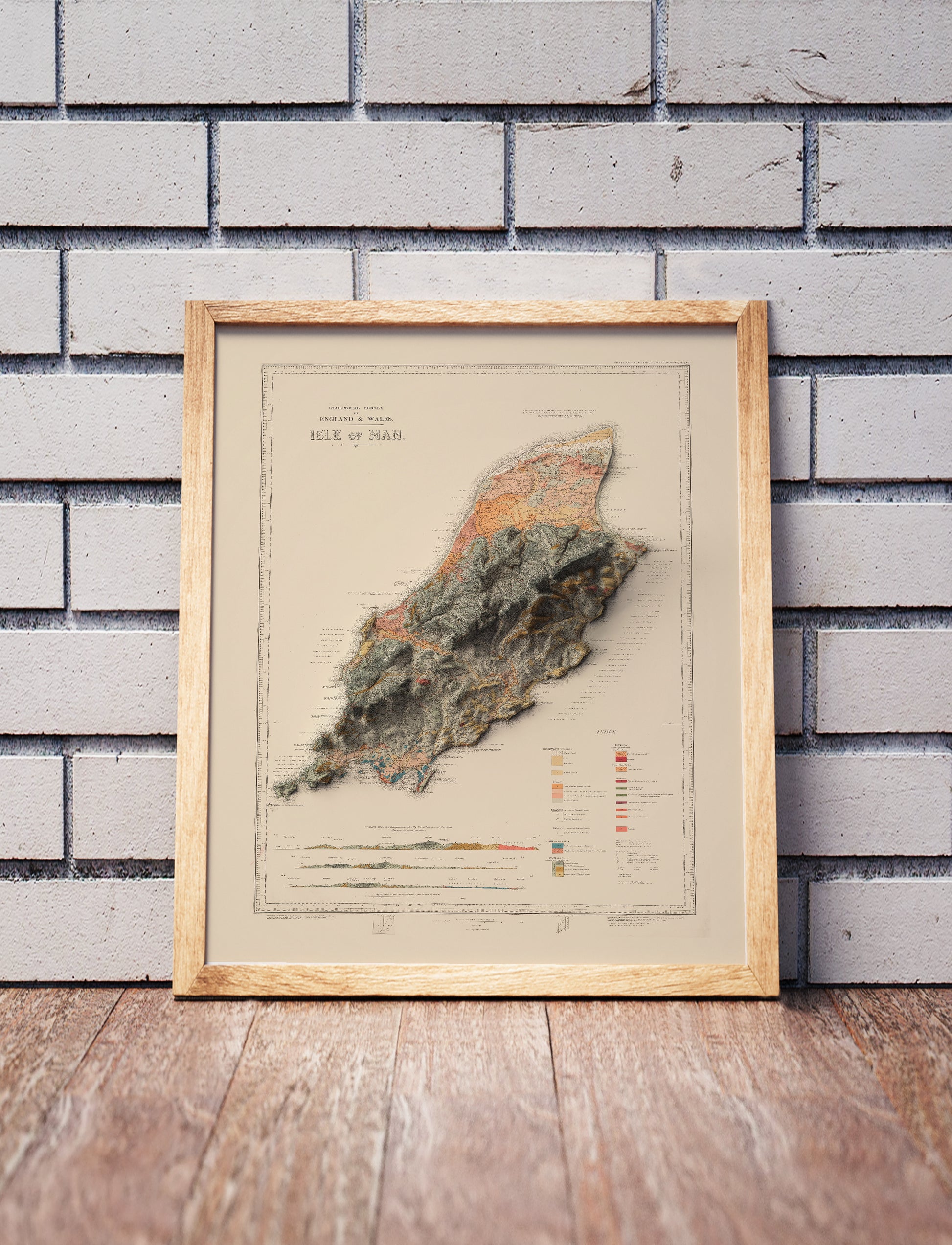 Vintage Isle of Man shaded relief map in a wooden frame. Realistic terrain art print for home decor, displayed against a brick wall