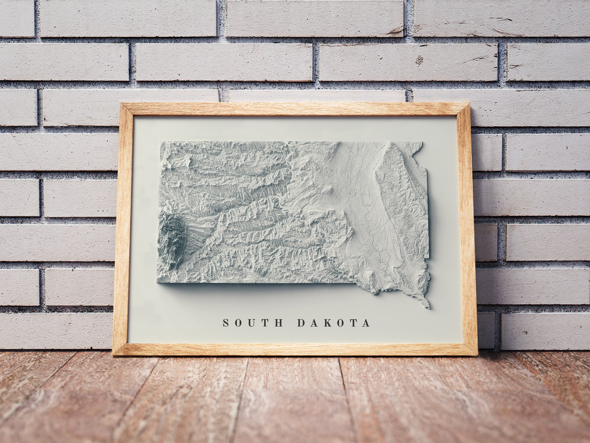 South Dakota shaded relief map in a wooden frame. Realistic terrain art print for home decor, displayed against a brick wall
