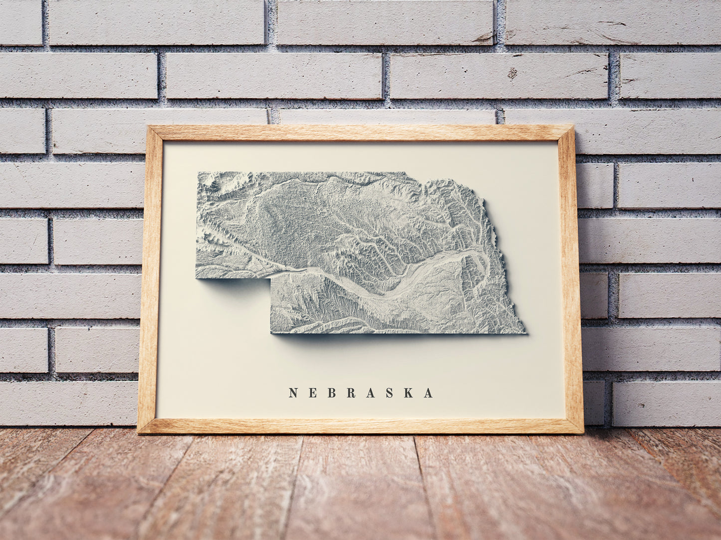 Nebraska shaded relief map in a wooden frame. Realistic terrain art print for home decor, displayed against a brick wall