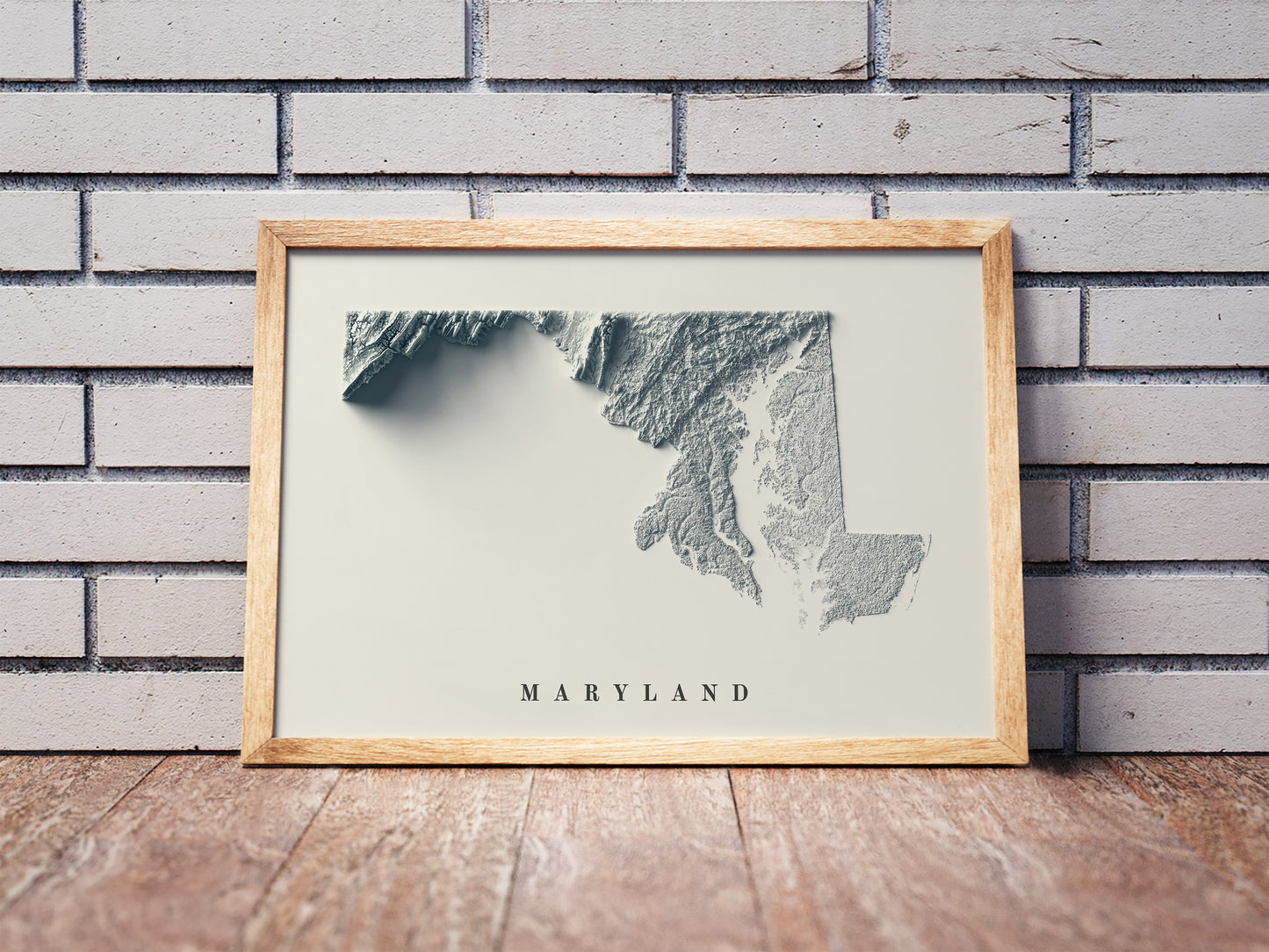 Maryland shaded relief map in a wooden frame. Realistic terrain art print for home decor, displayed against a brick wall