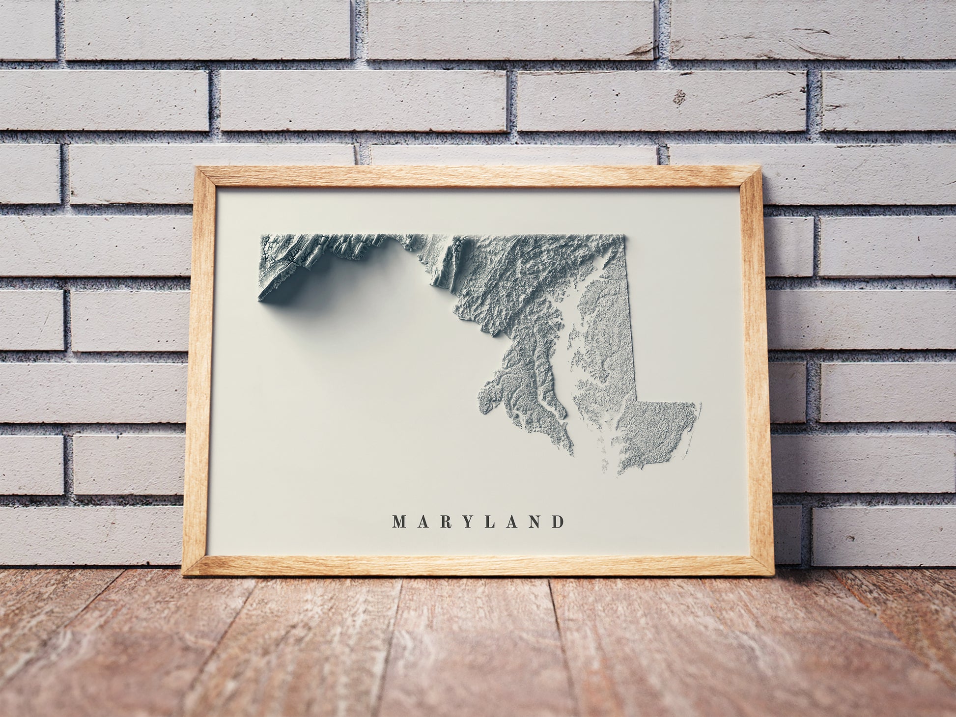 Maryland shaded relief map in a wooden frame. Realistic terrain art print for home decor, displayed against a brick wall