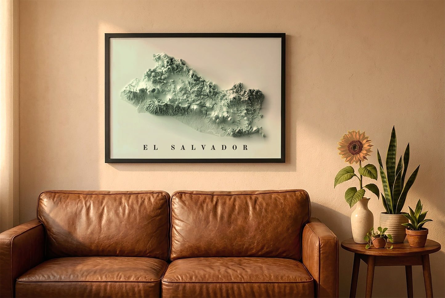 El Salvador relief map in a black frame above a brown leather sofa. Boho living room with warm sunlight, sunflower, and indoor plants on a wooden side table