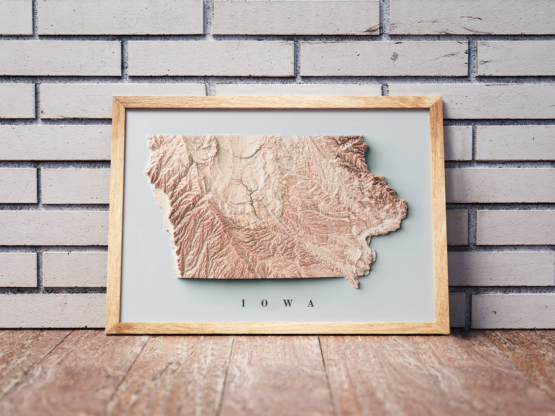 Iowa shaded relief map in a wooden frame. Realistic terrain art print for home decor, displayed against a brick wall