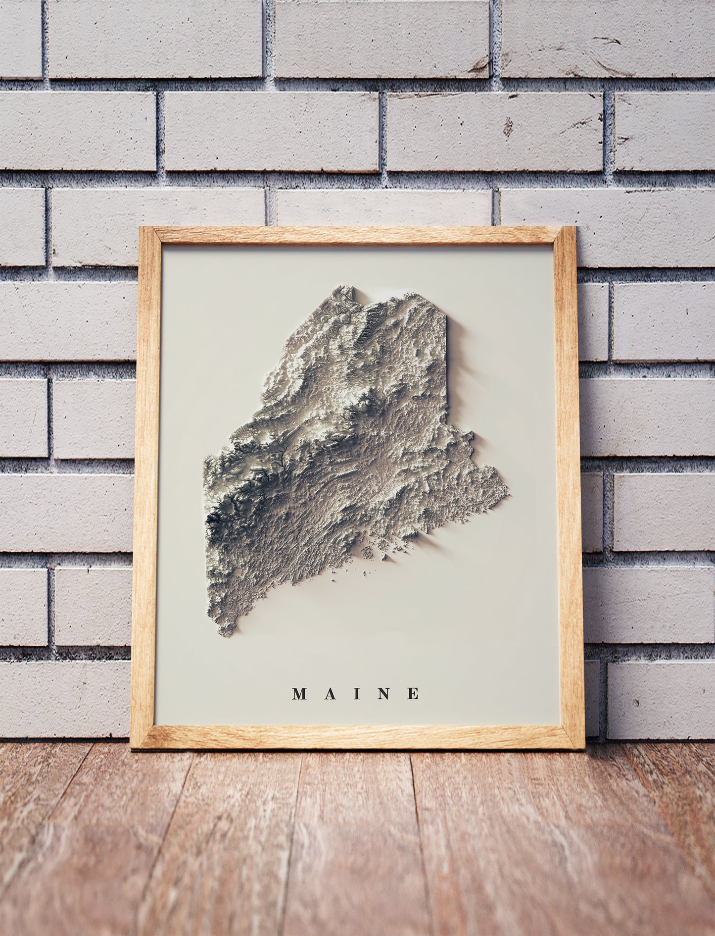 Maine shaded relief map in a wooden frame. Realistic terrain art print for home decor, displayed against a brick wall