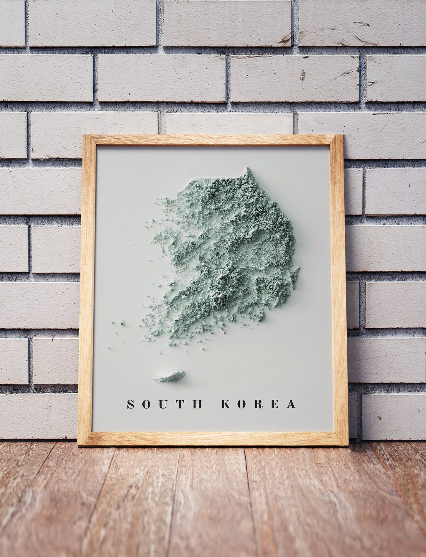 South Korea shaded relief map in a wooden frame. Realistic terrain art print for home decor, displayed against a brick wall