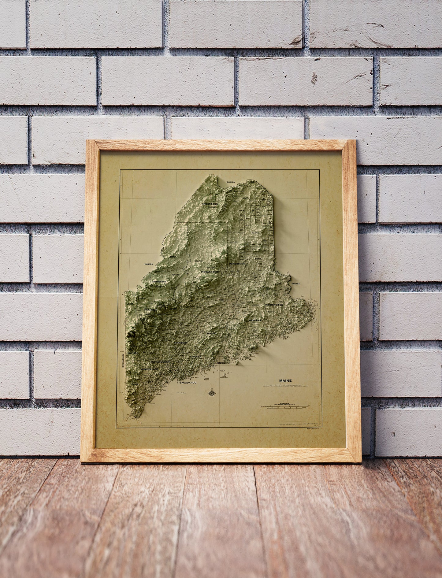 Vintage Maine shaded relief map in a wooden frame. Realistic terrain art print for home decor, displayed against a brick wall