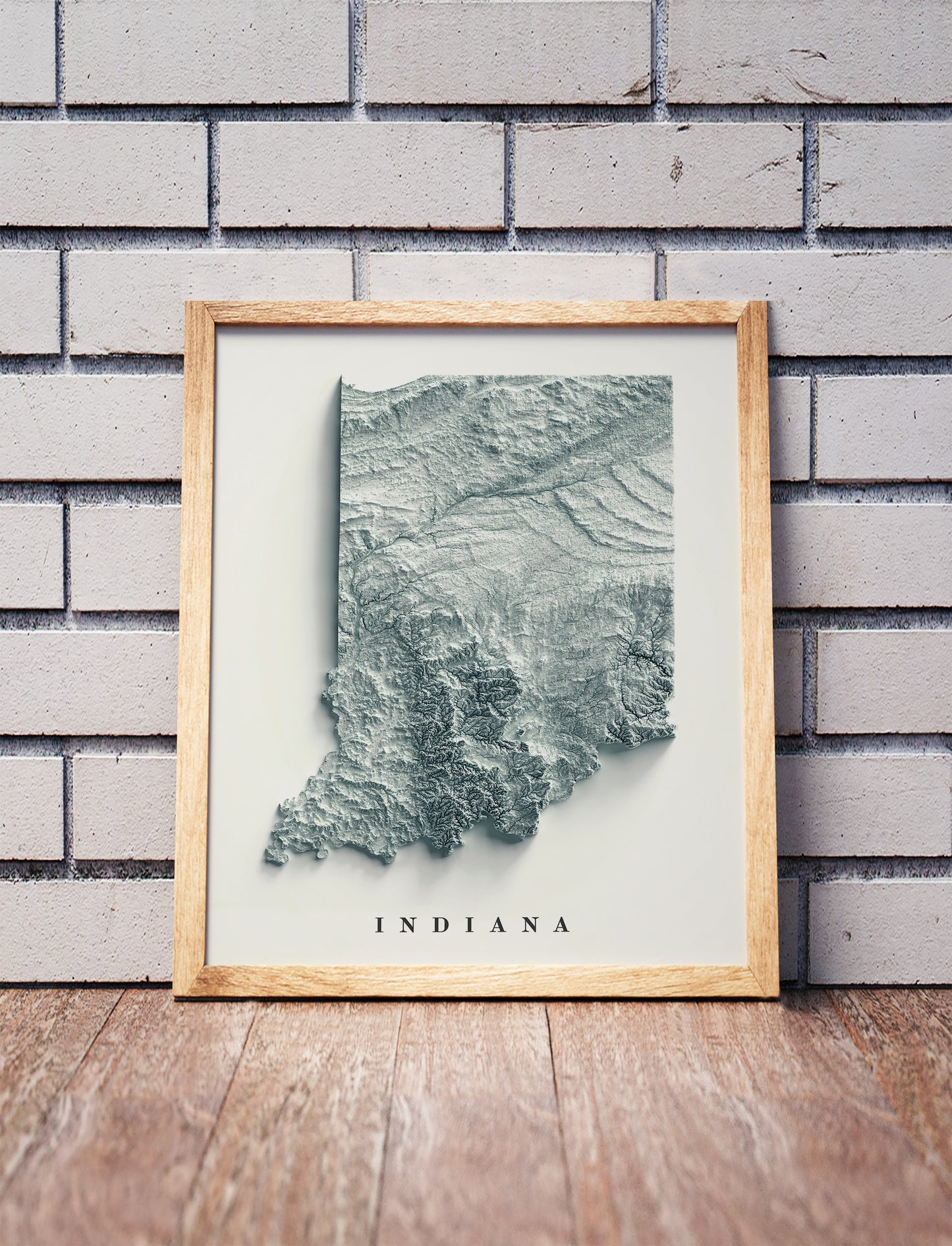 Indiana shaded relief map in a wooden frame. Realistic terrain art print for home decor, displayed against a brick wall