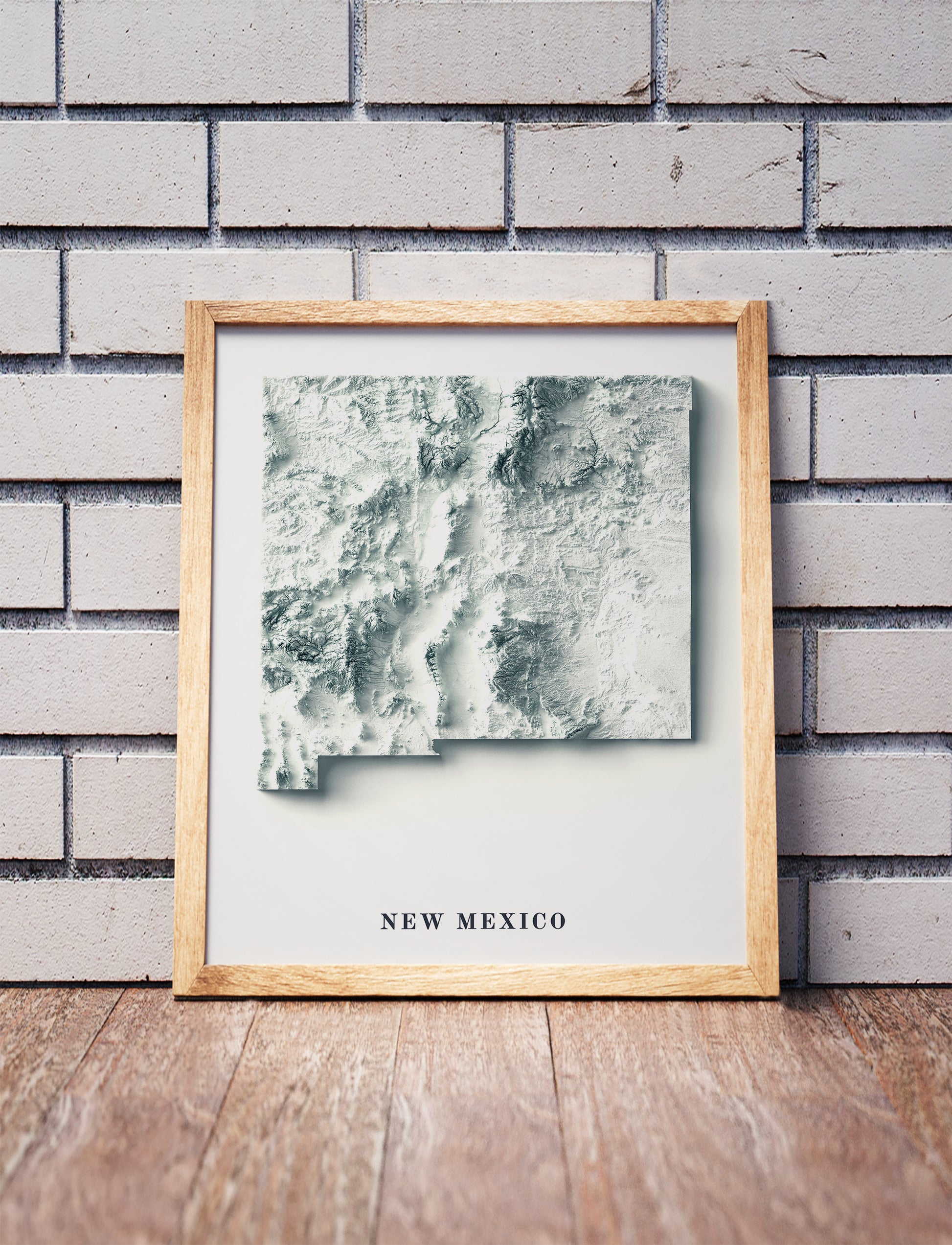 New Mexico shaded relief map in a wooden frame. Realistic terrain art print for home decor, displayed against a brick wall