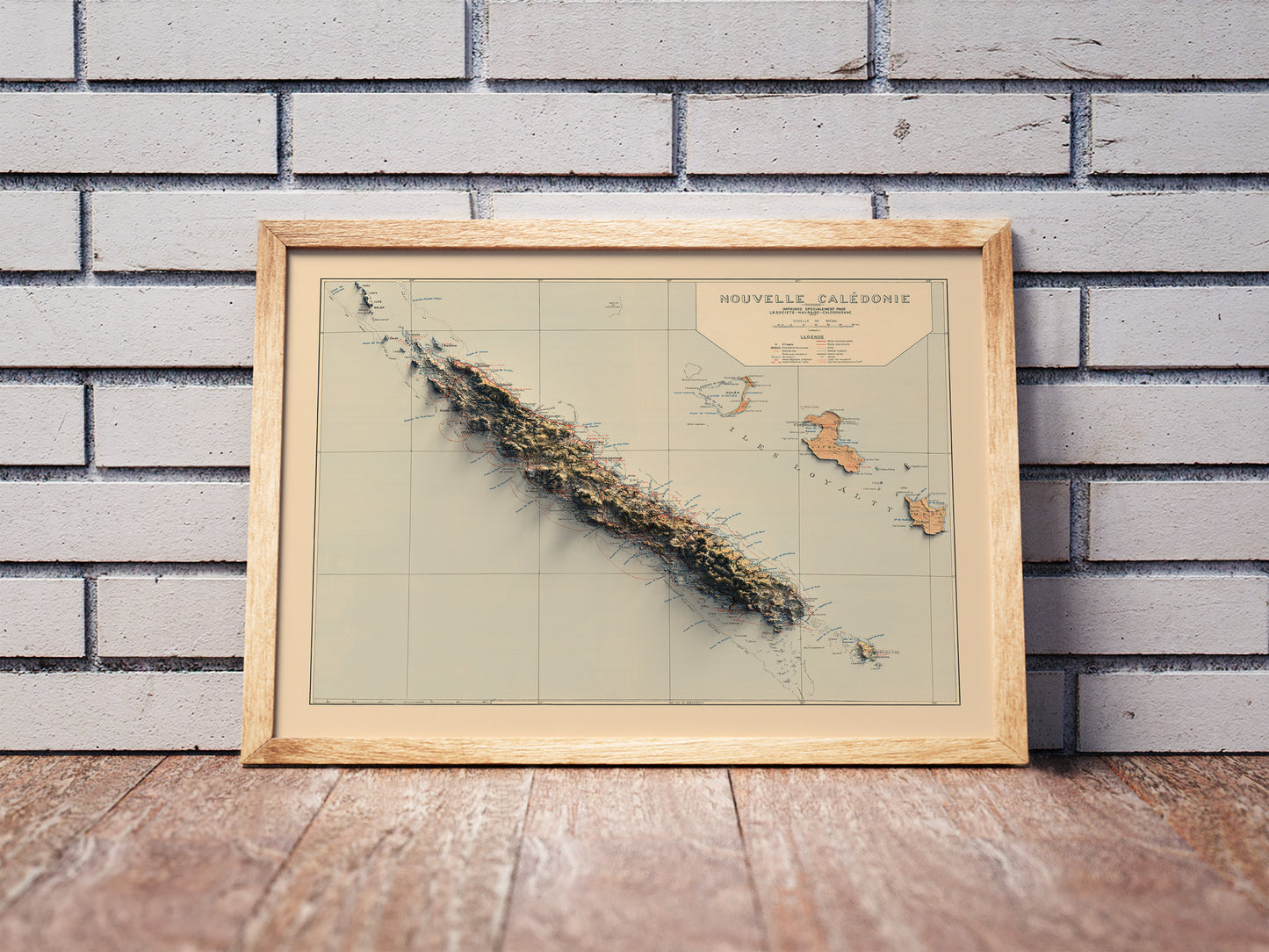 Vintage New Caledonia shaded relief map in a wooden frame. Realistic terrain art print for home decor, displayed against a brick wall