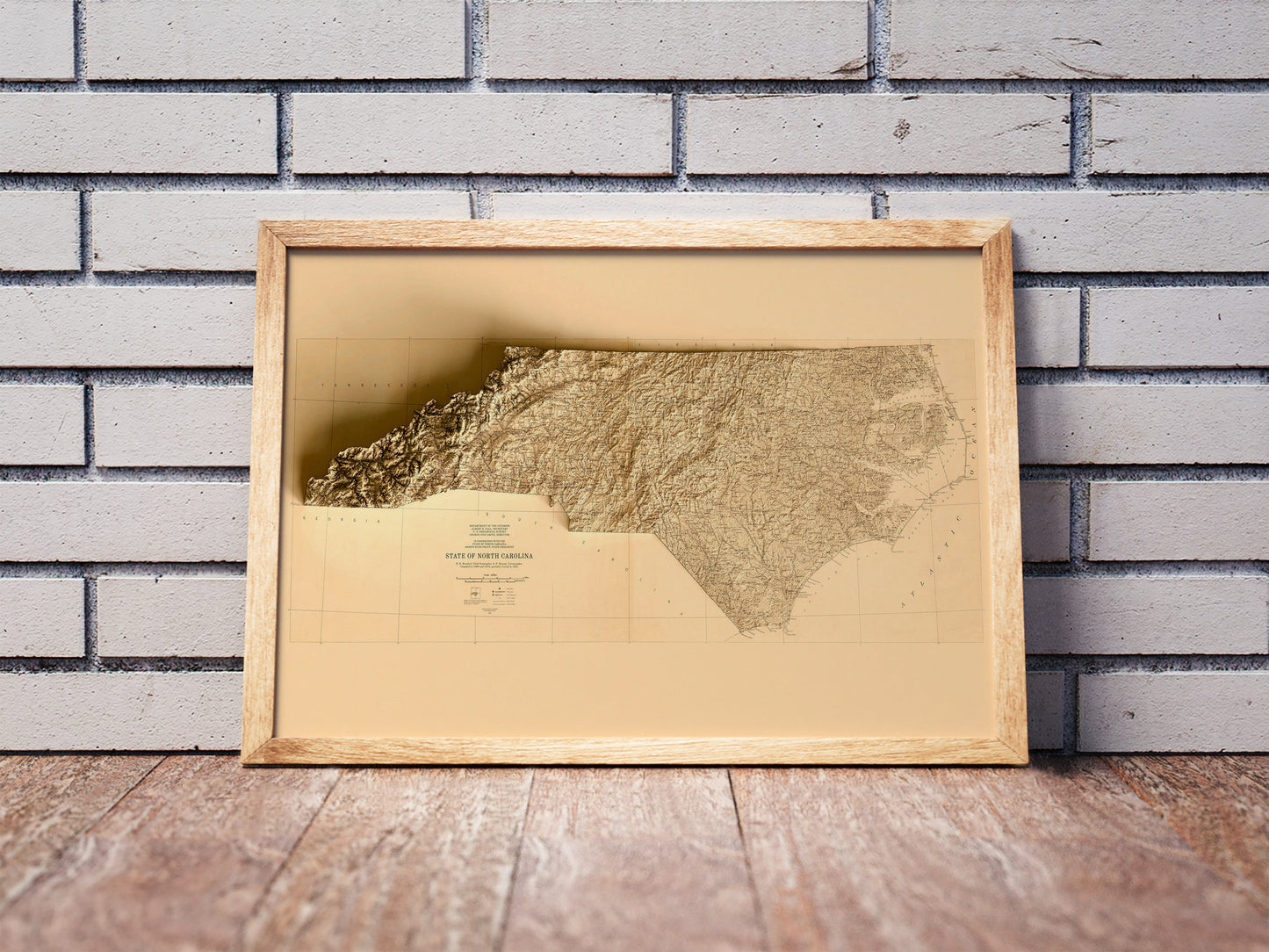 Vintage North Carolina shaded relief map in a wooden frame. Realistic terrain art print for home decor, displayed against a brick wall