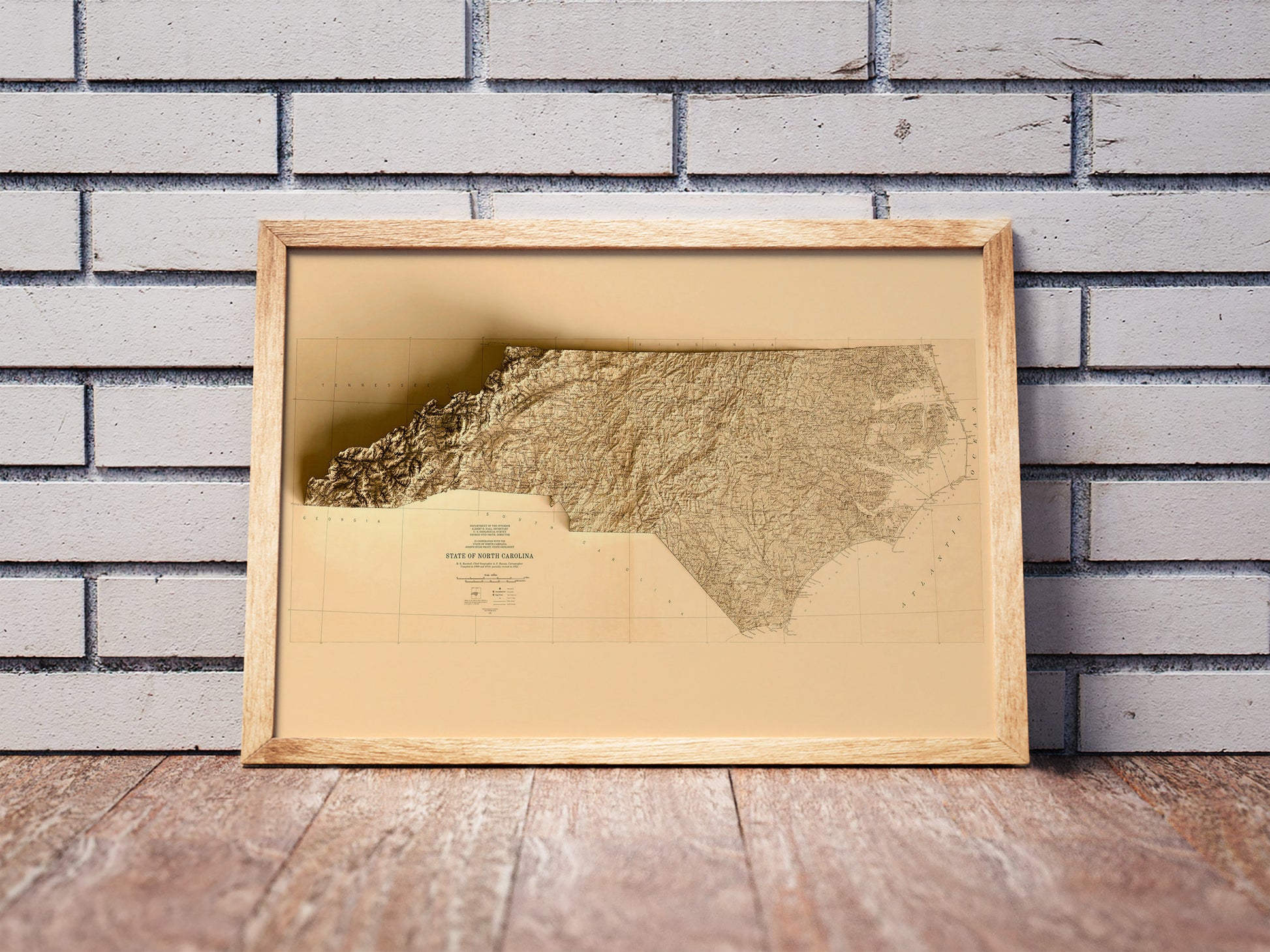 Vintage North Carolina shaded relief map in a wooden frame. Realistic terrain art print for home decor, displayed against a brick wall