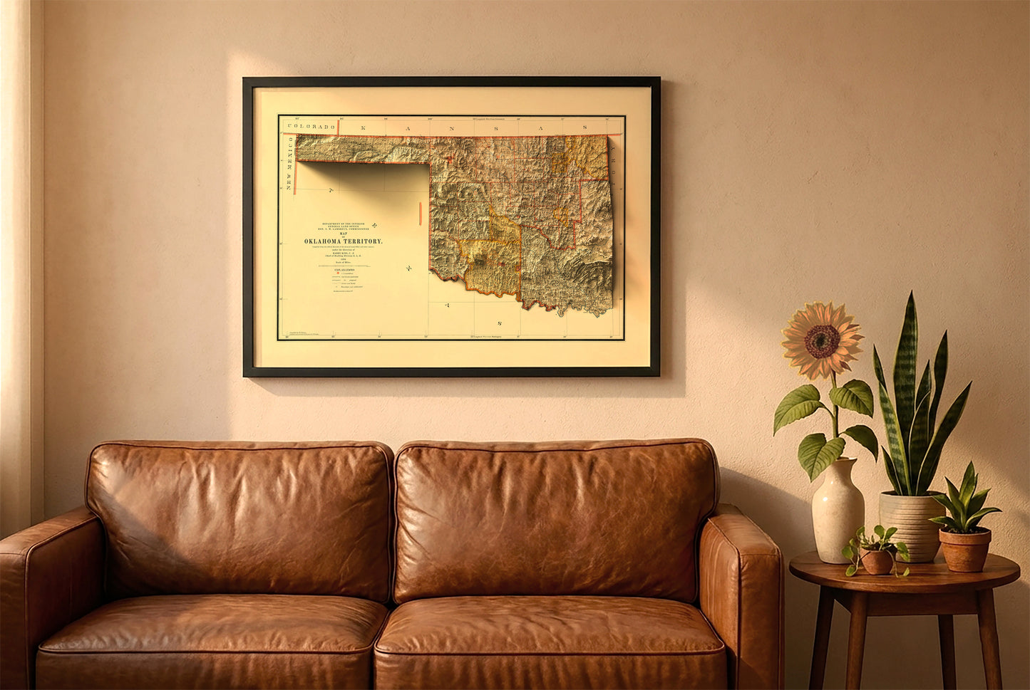 Oklahoma vintage relief map in a black frame above a brown leather sofa. Boho living room with warm sunlight, sunflower, and indoor plants on a wooden side table