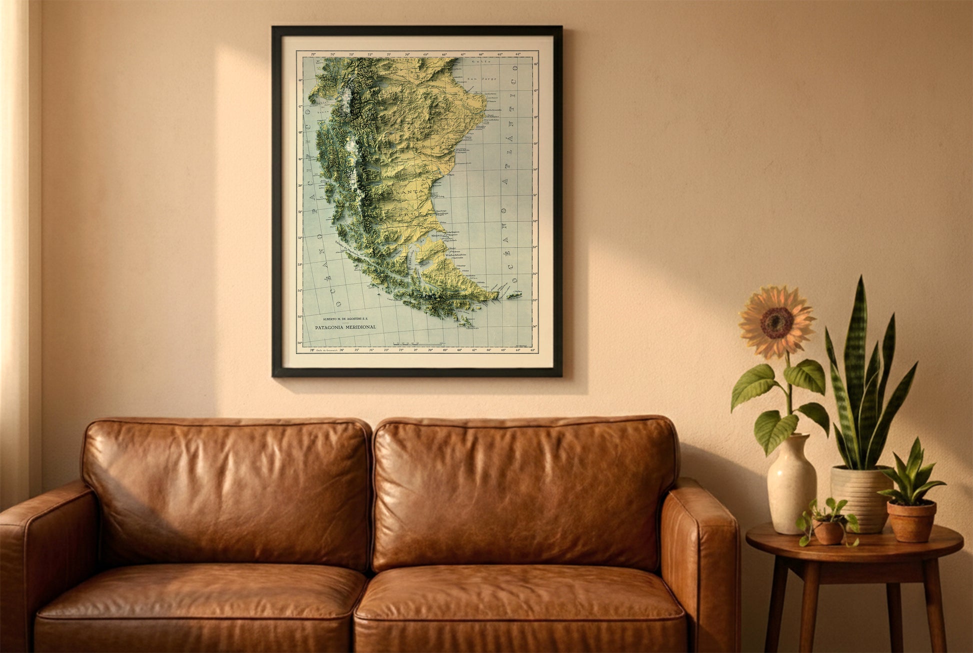 Patagonia vintage relief map in a black frame above a brown leather sofa. Boho living room with warm sunlight, sunflower, and indoor plants on a wooden side table