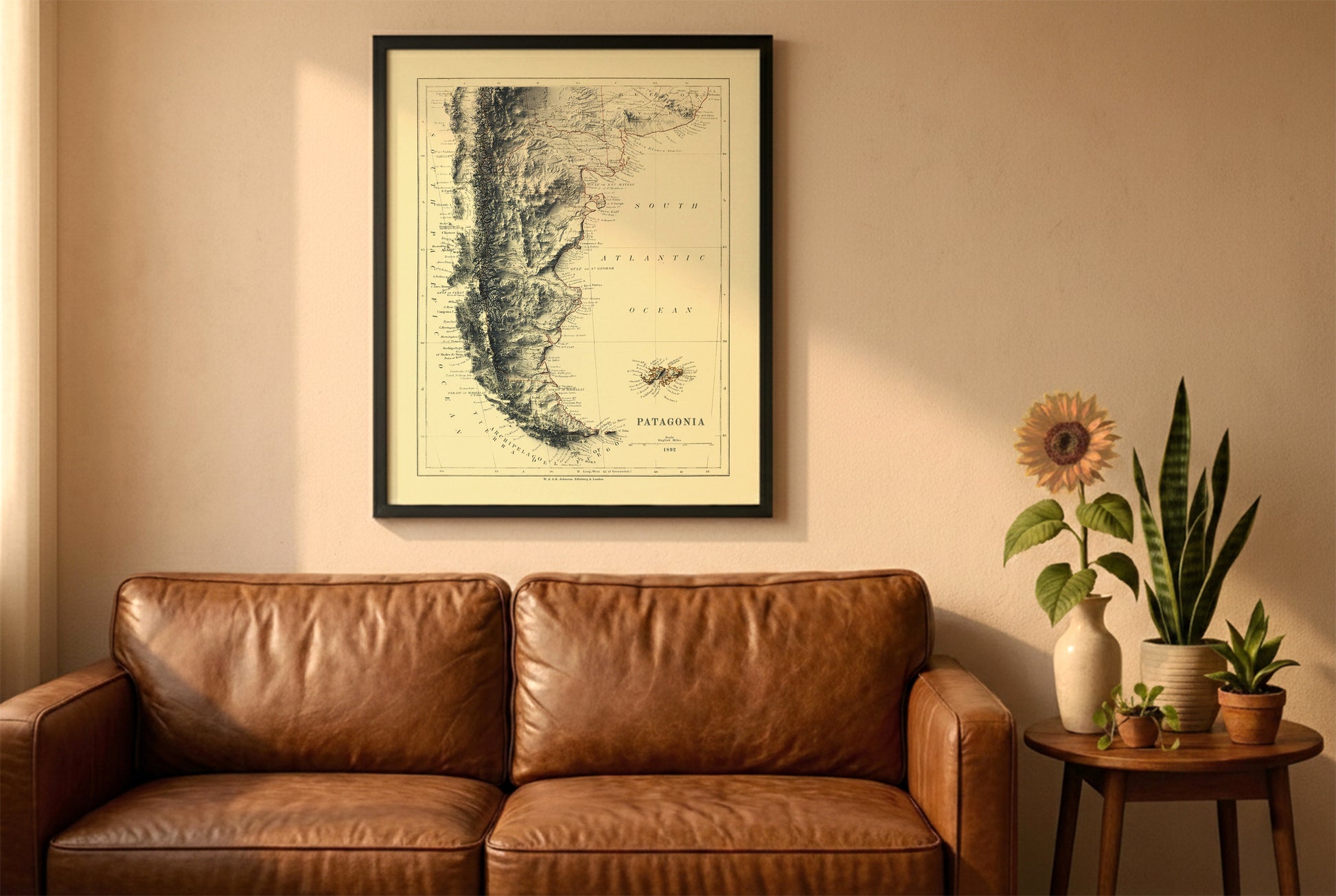 Patagonia vintage relief map in a black frame above a brown leather sofa. Boho living room with warm sunlight, sunflower, and indoor plants on a wooden side table