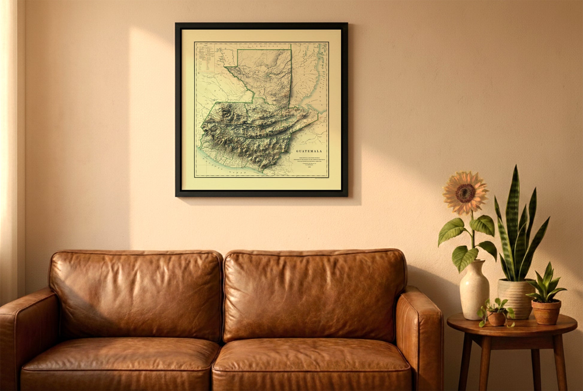 Guatemala vintage relief map in a black frame above a brown leather sofa. Boho living room with warm sunlight, sunflower, and indoor plants on a wooden side table