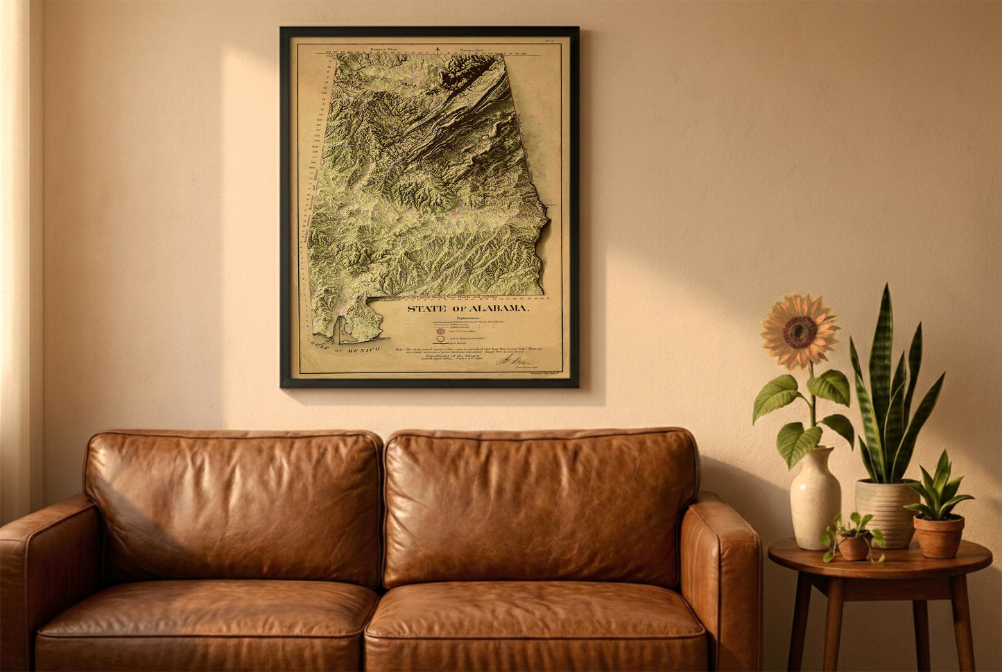 Alabama vintage relief map in a black frame above a brown leather sofa. Boho living room with warm sunlight, sunflower, and indoor plants on a wooden side table