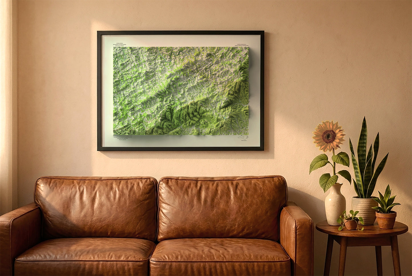 Alleghany County vintage relief map in a black frame above a brown leather sofa. Boho living room with warm sunlight, sunflower, and indoor plants on a wooden side table
