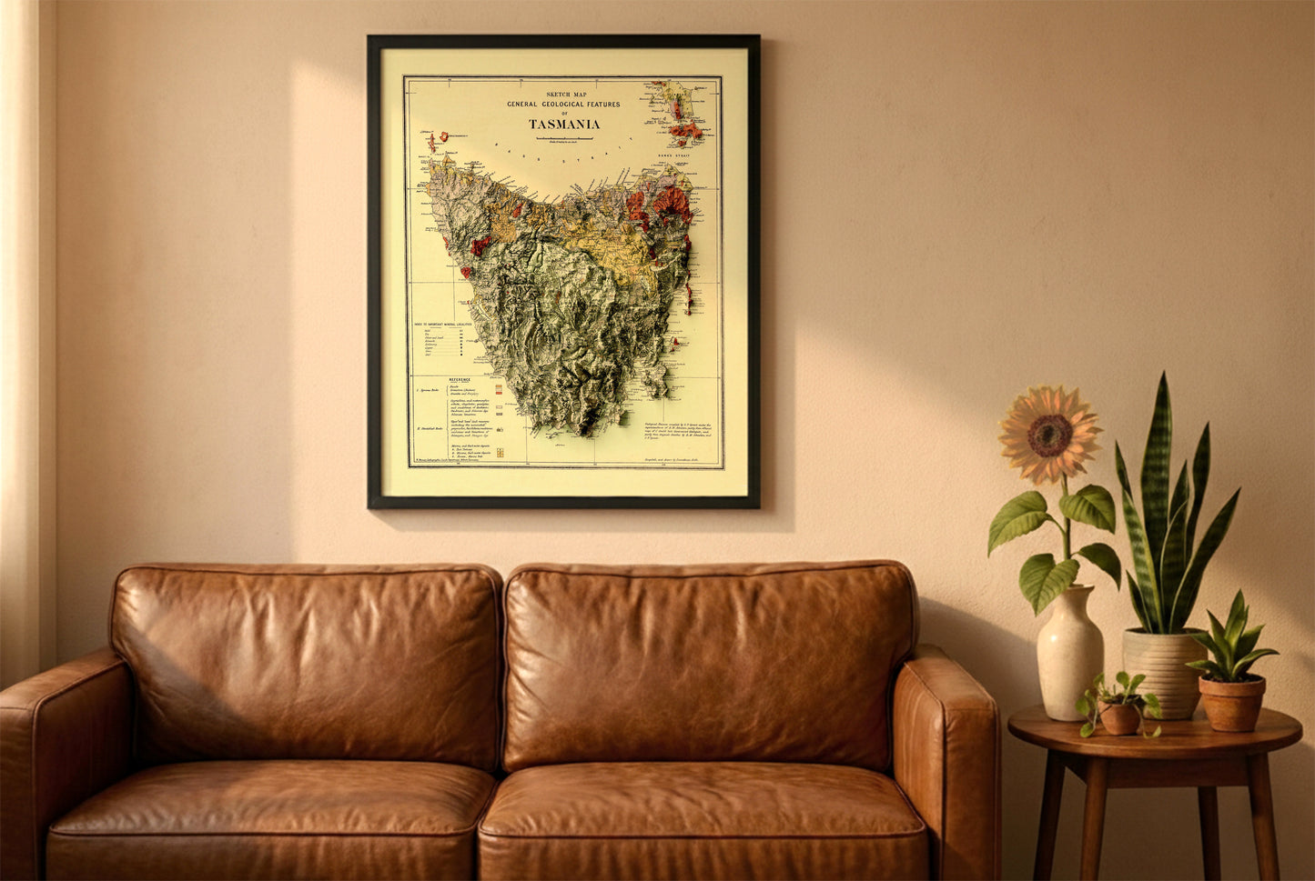 Tasmania vintage relief map in a black frame above a brown leather sofa. Boho living room with warm sunlight, sunflower, and indoor plants on a wooden side table