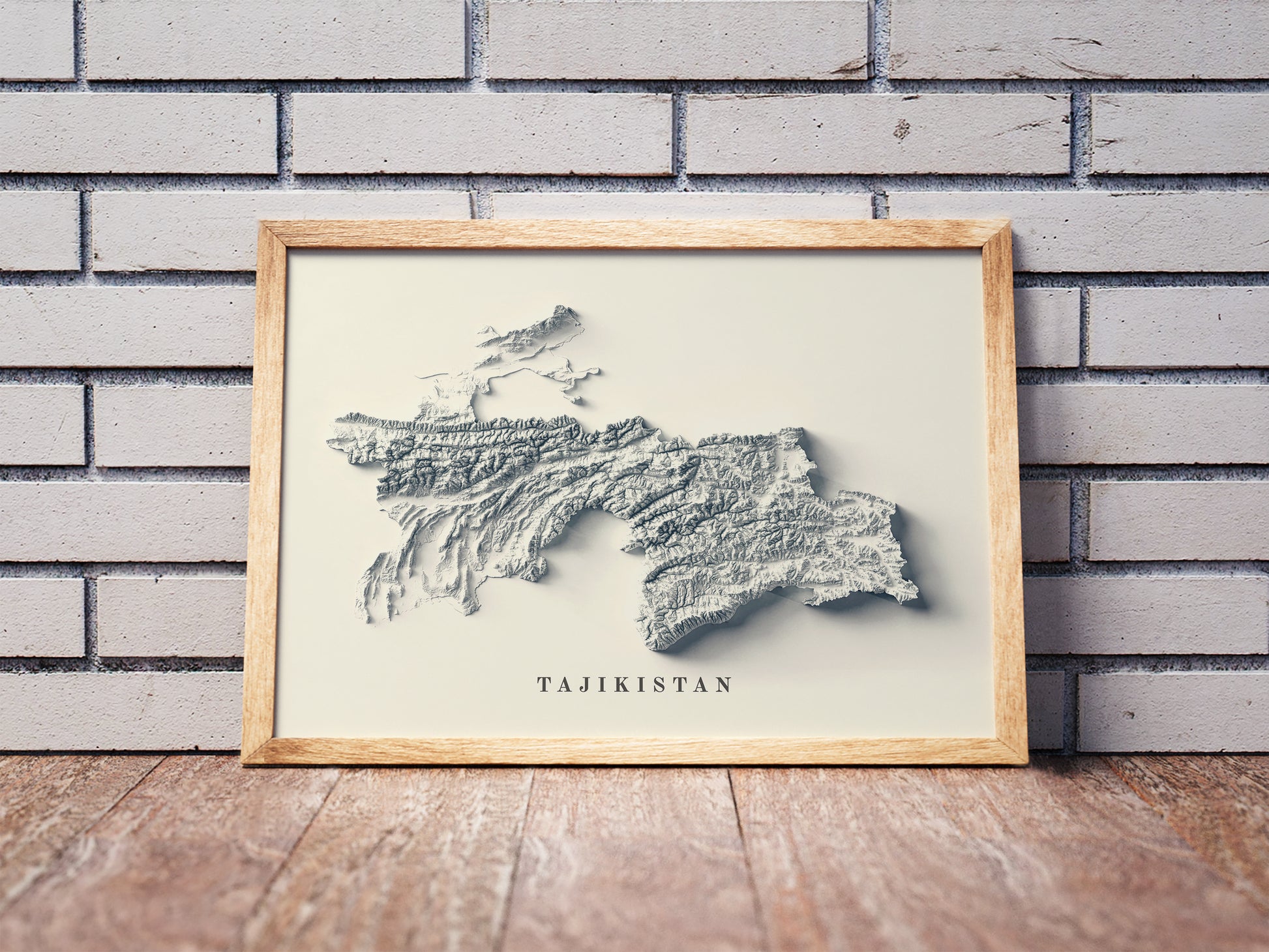 Tajikistan shaded relief map in a wooden frame. Realistic terrain art print for home decor, displayed against a brick wall