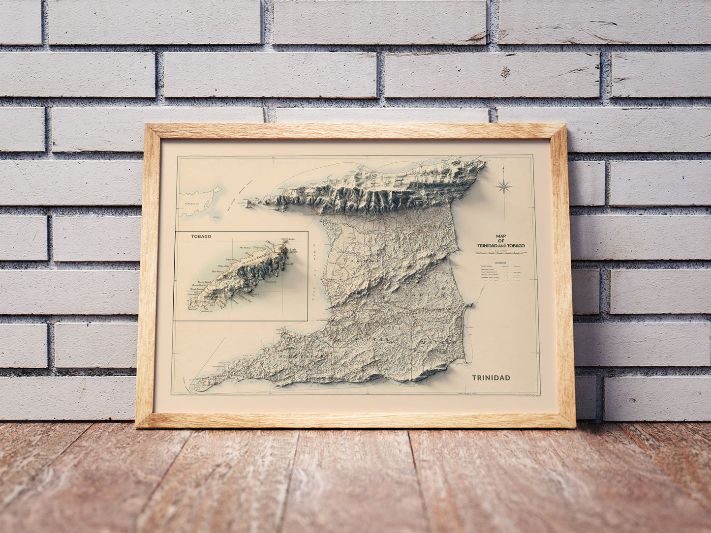 Vintage Trinidad and Tobago shaded relief map in a wooden frame. Realistic terrain art print for home decor, displayed against a brick wall