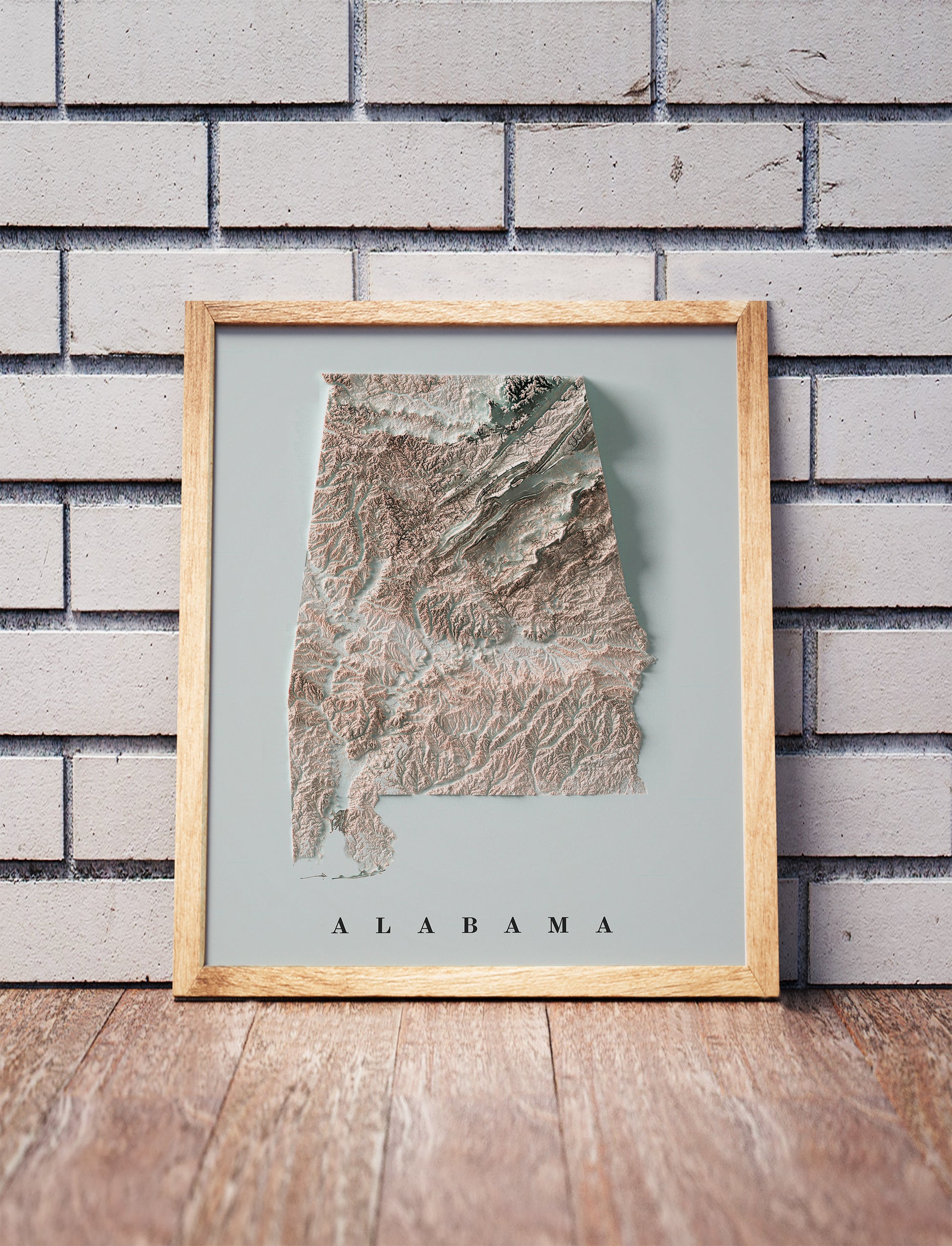 Alabama shaded relief map in a wooden frame. Realistic terrain art print for home decor, displayed against a brick wall