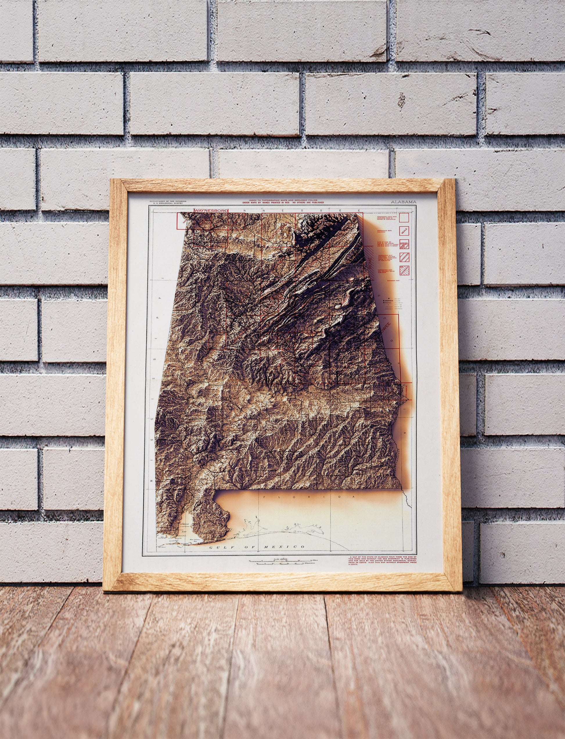 Vintage Alabama shaded relief map in a wooden frame. Realistic terrain art print for home decor, displayed against a brick wall
