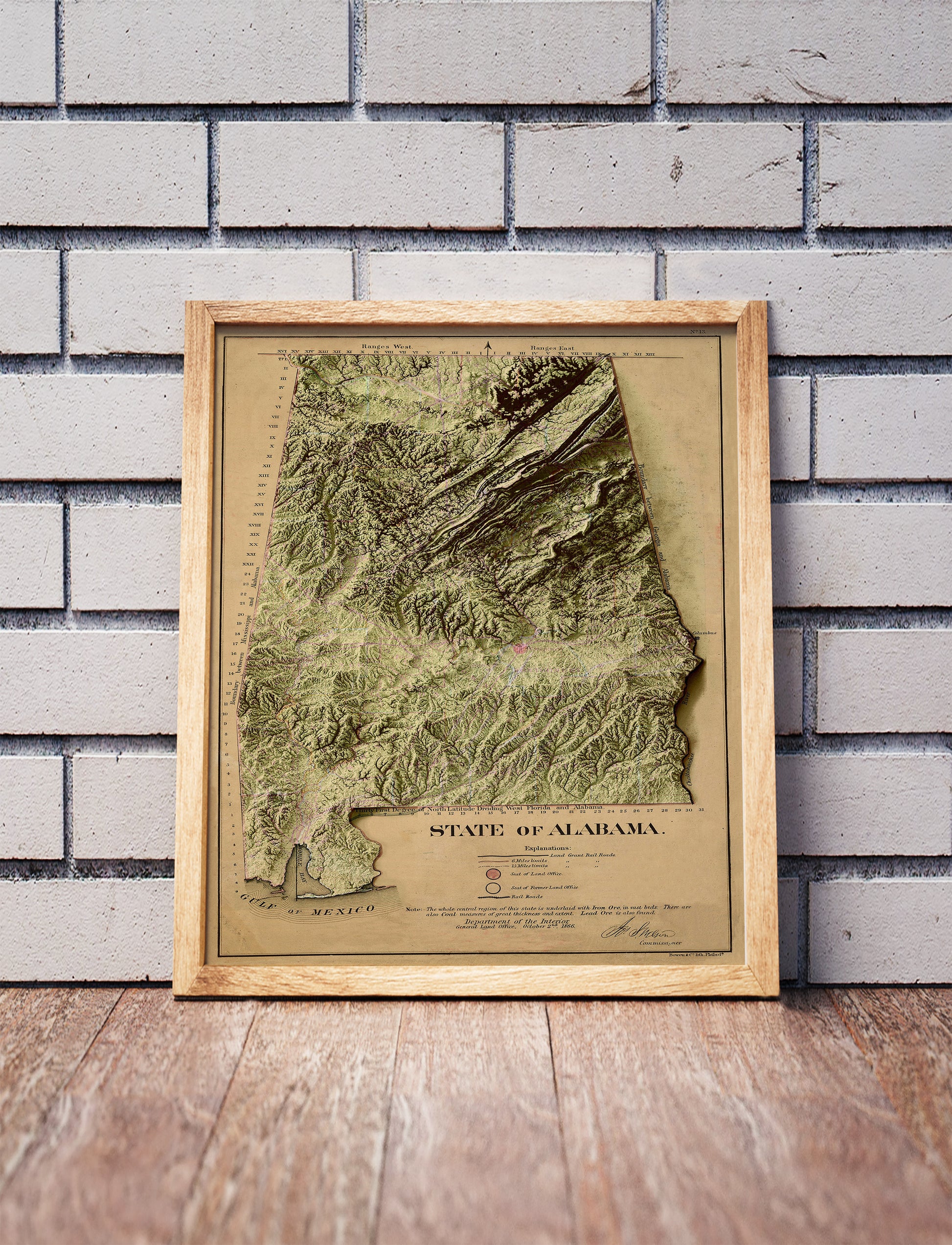 Vintage Alabama shaded relief map in a wooden frame. Realistic terrain art print for home decor, displayed against a brick wall