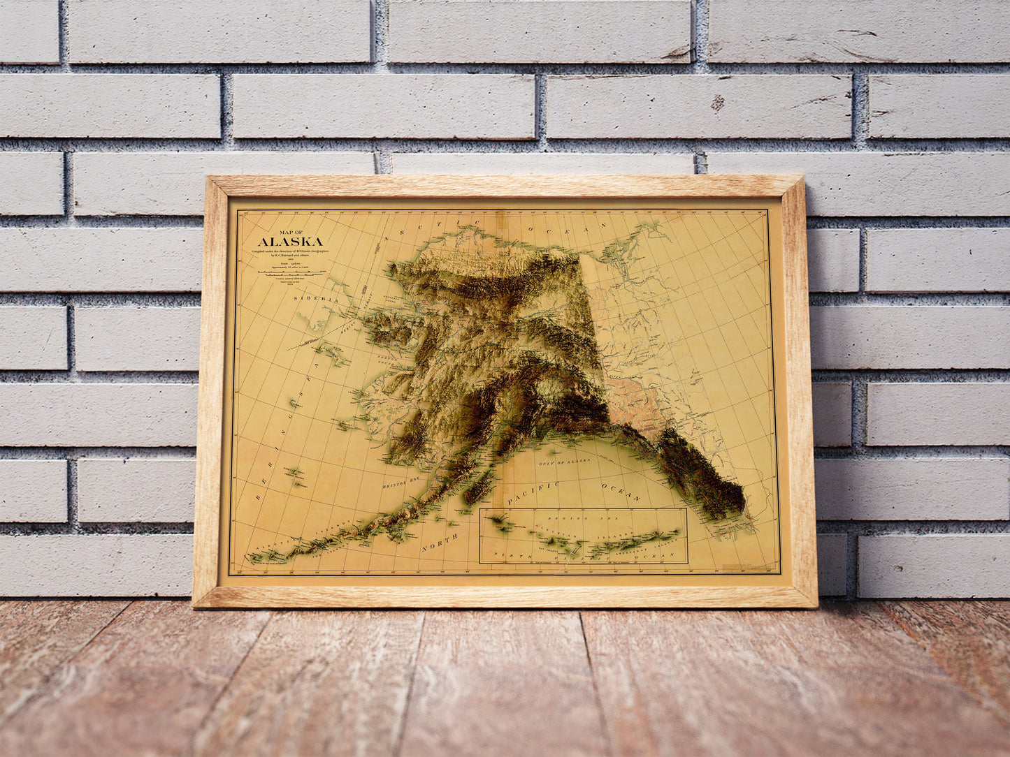 Vintage Alaska shaded relief map in a wooden frame. Realistic terrain art print for home decor, displayed against a brick wall