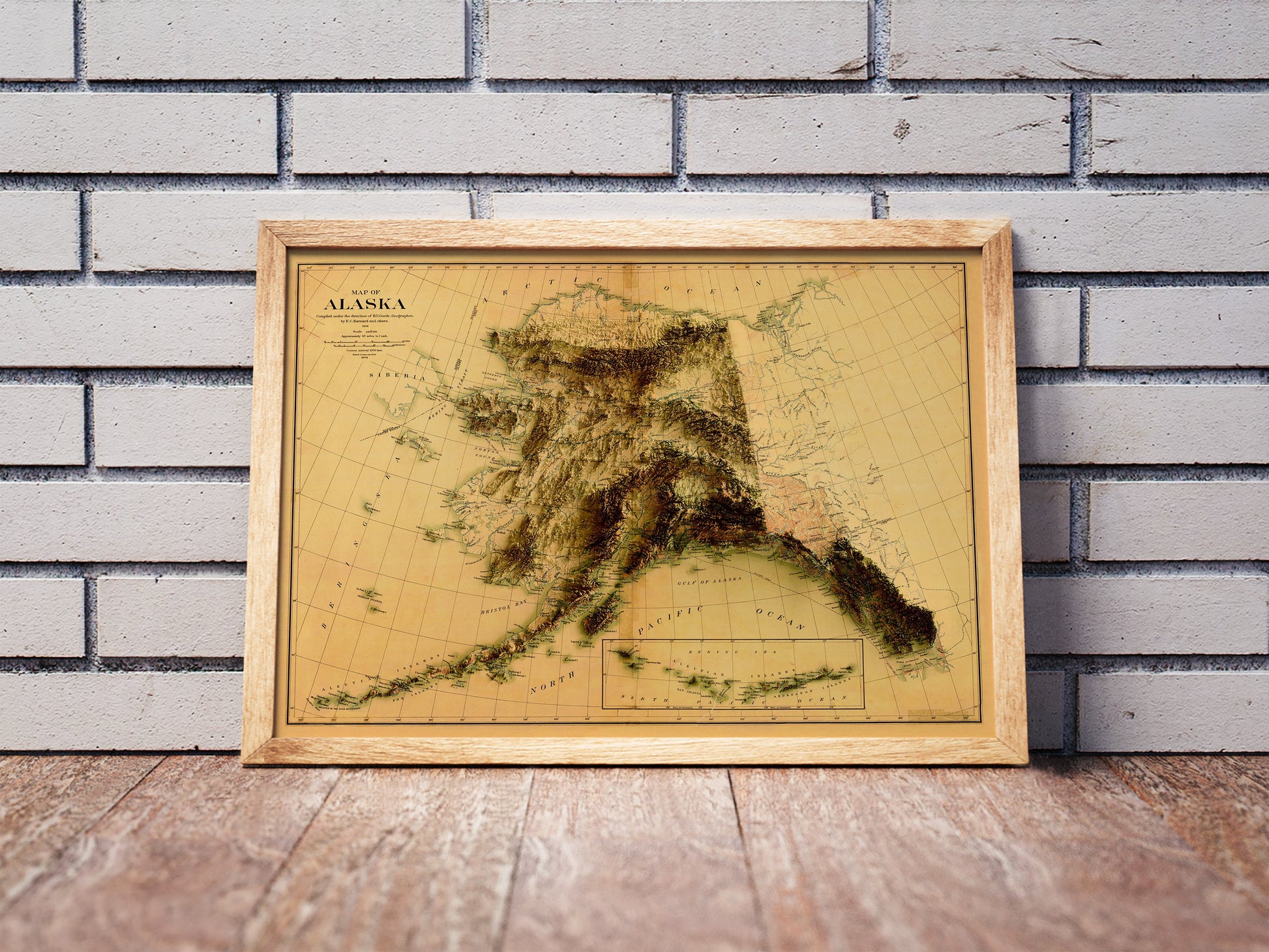Vintage Alaska shaded relief map in a wooden frame. Realistic terrain art print for home decor, displayed against a brick wall