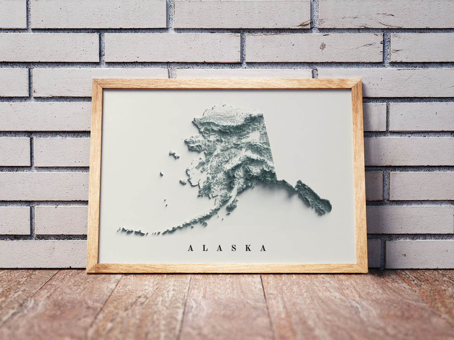 Alaska shaded relief map in a wooden frame. Realistic terrain art print for home decor, displayed against a brick wall