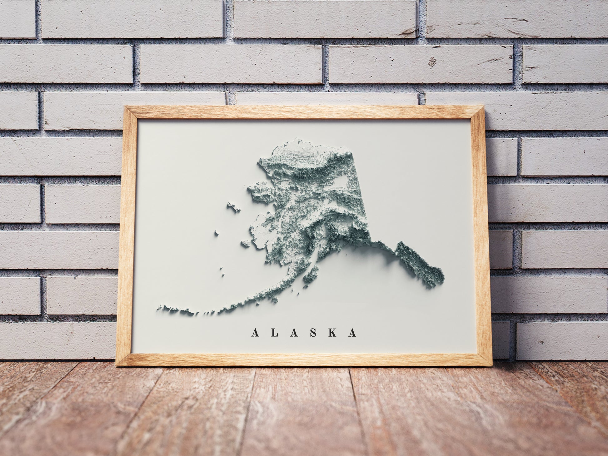 Alaska shaded relief map in a wooden frame. Realistic terrain art print for home decor, displayed against a brick wall