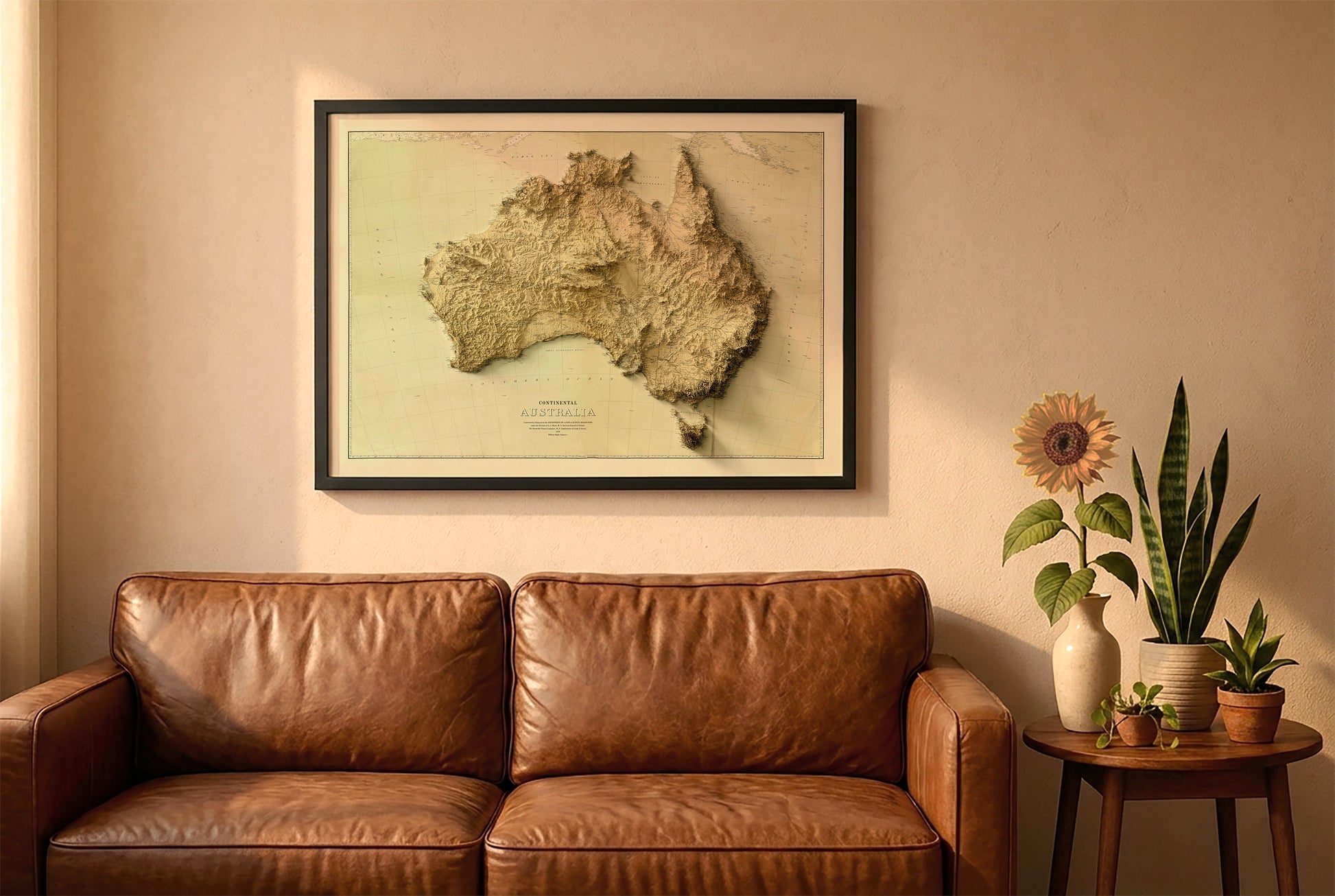 Australia vintage relief map in a black frame above a brown leather sofa. Boho living room with warm sunlight, sunflower, and indoor plants on a wooden side table