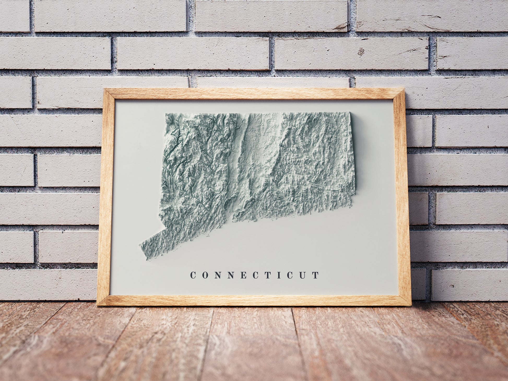 Connecticut shaded relief map in a wooden frame. Realistic terrain art print for home decor, displayed against a brick wall