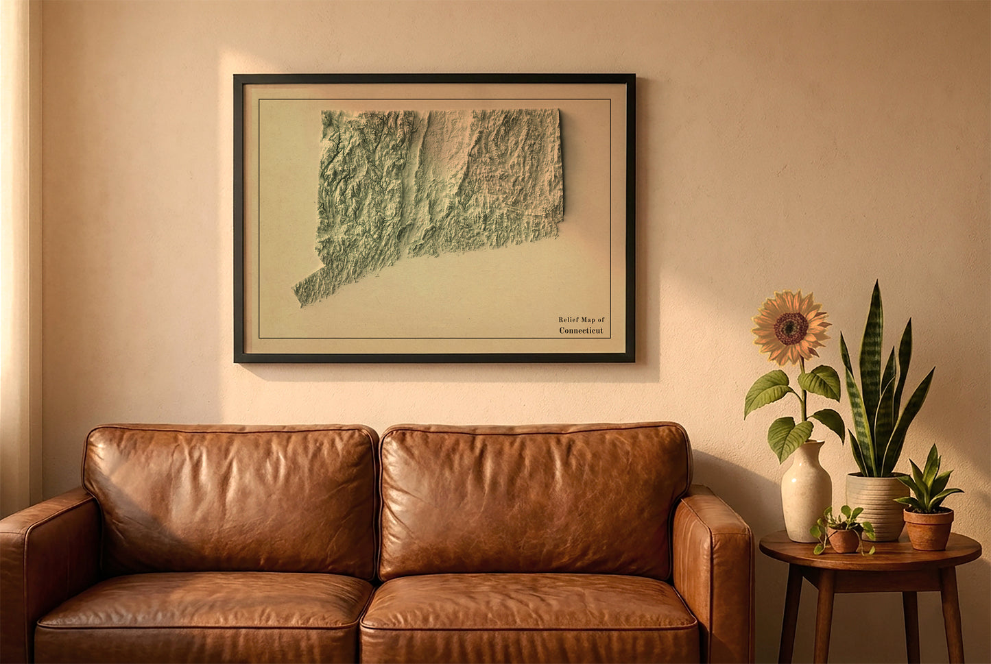 Connecticut vintage relief map in a black frame above a brown leather sofa. Boho living room with warm sunlight, sunflower, and indoor plants on a wooden side table