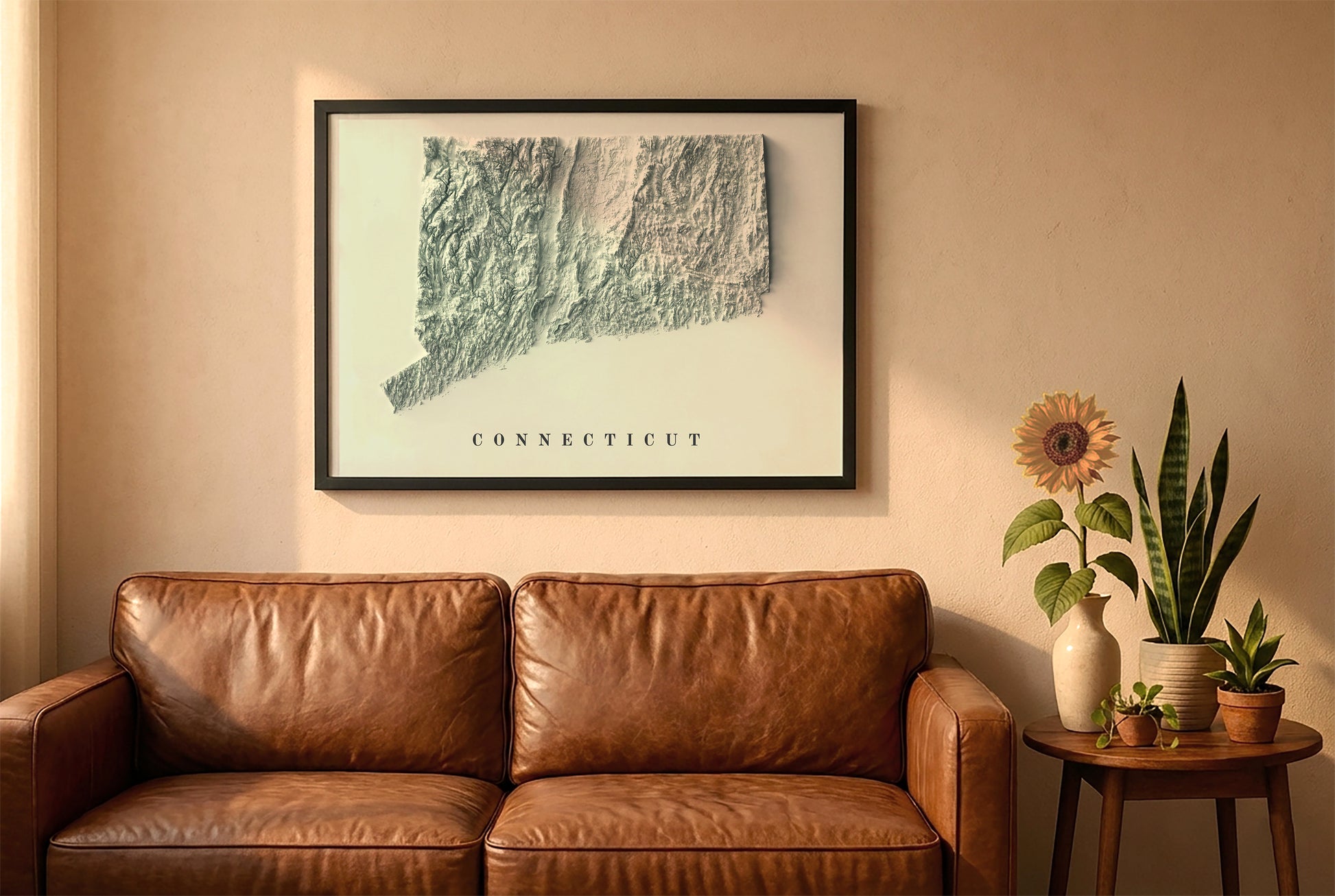 Connecticut relief map in a black frame above a brown leather sofa. Boho living room with warm sunlight, sunflower, and indoor plants on a wooden side table
