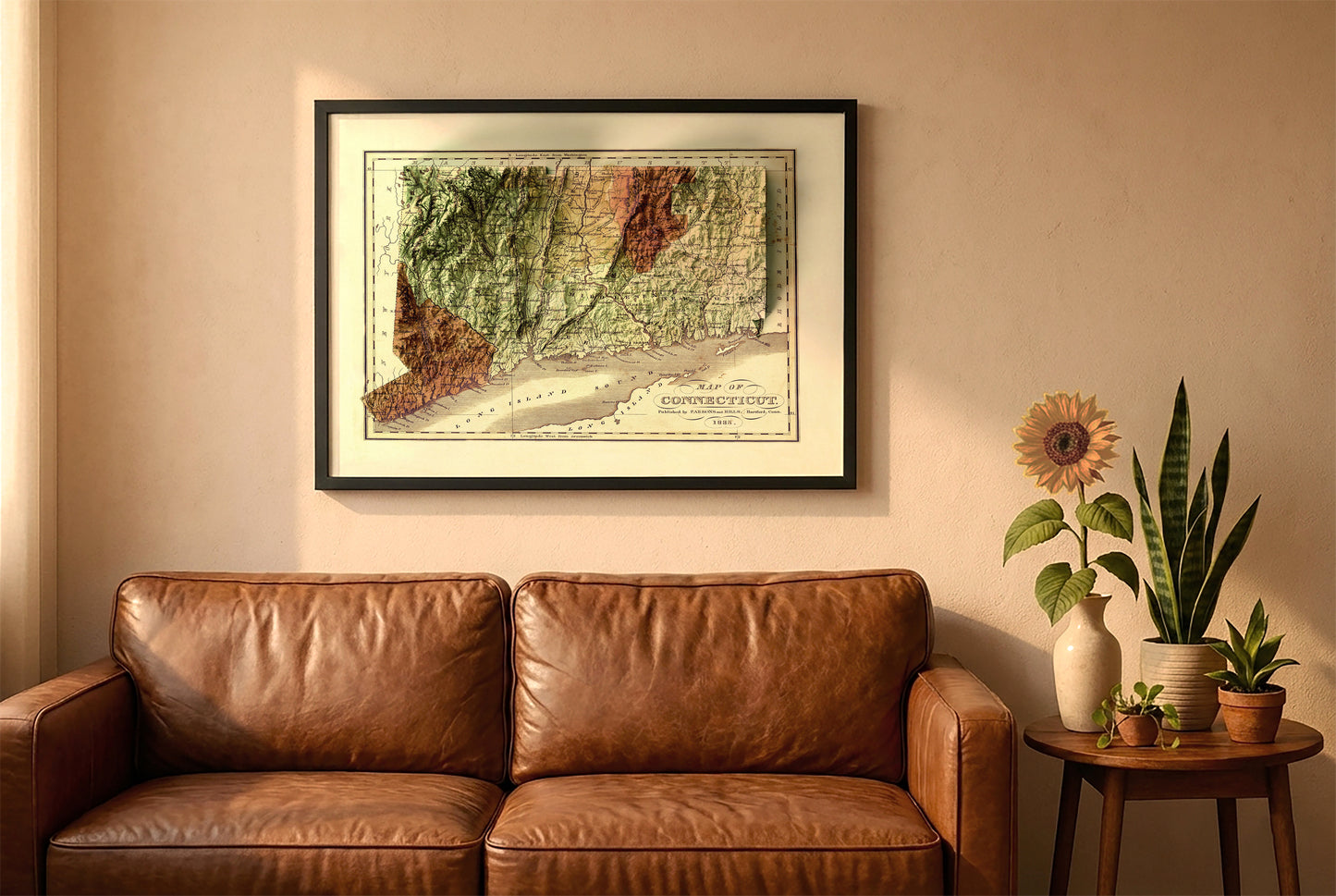 Connecticut vintage relief map in a black frame above a brown leather sofa. Boho living room with warm sunlight, sunflower, and indoor plants on a wooden side table