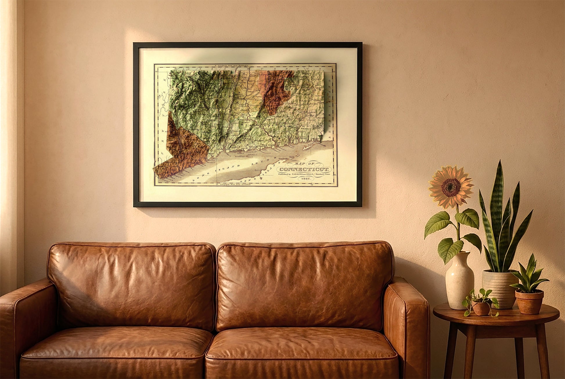 Connecticut vintage relief map in a black frame above a brown leather sofa. Boho living room with warm sunlight, sunflower, and indoor plants on a wooden side table