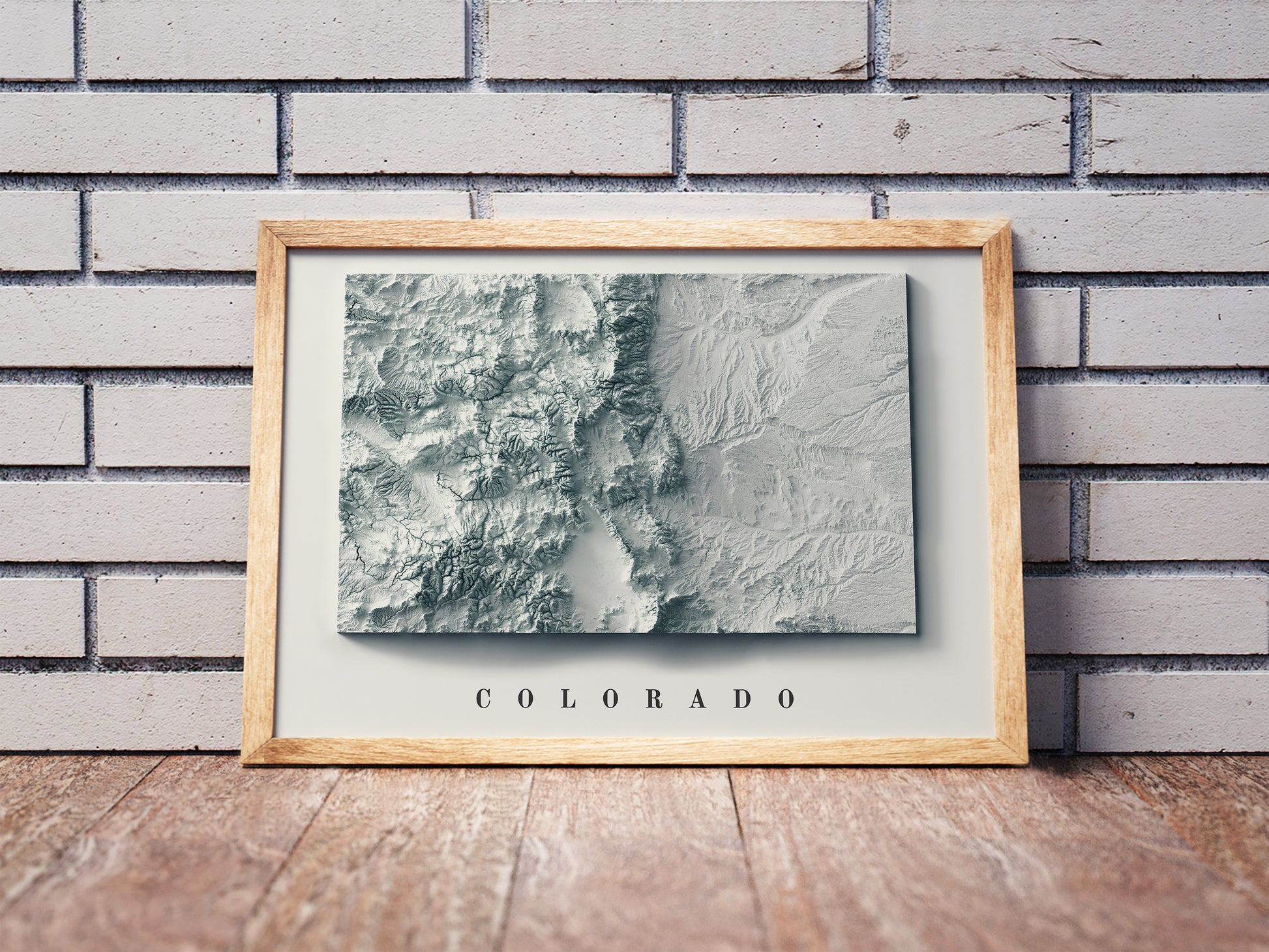 Colorado shaded relief map in a wooden frame. Realistic terrain art print for home decor, displayed against a brick wall