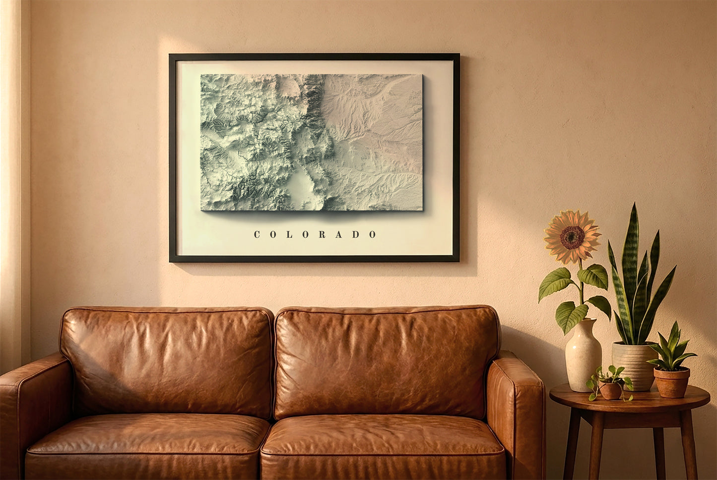 Colorado relief map in a black frame above a brown leather sofa. Boho living room with warm sunlight, sunflower, and indoor plants on a wooden side table