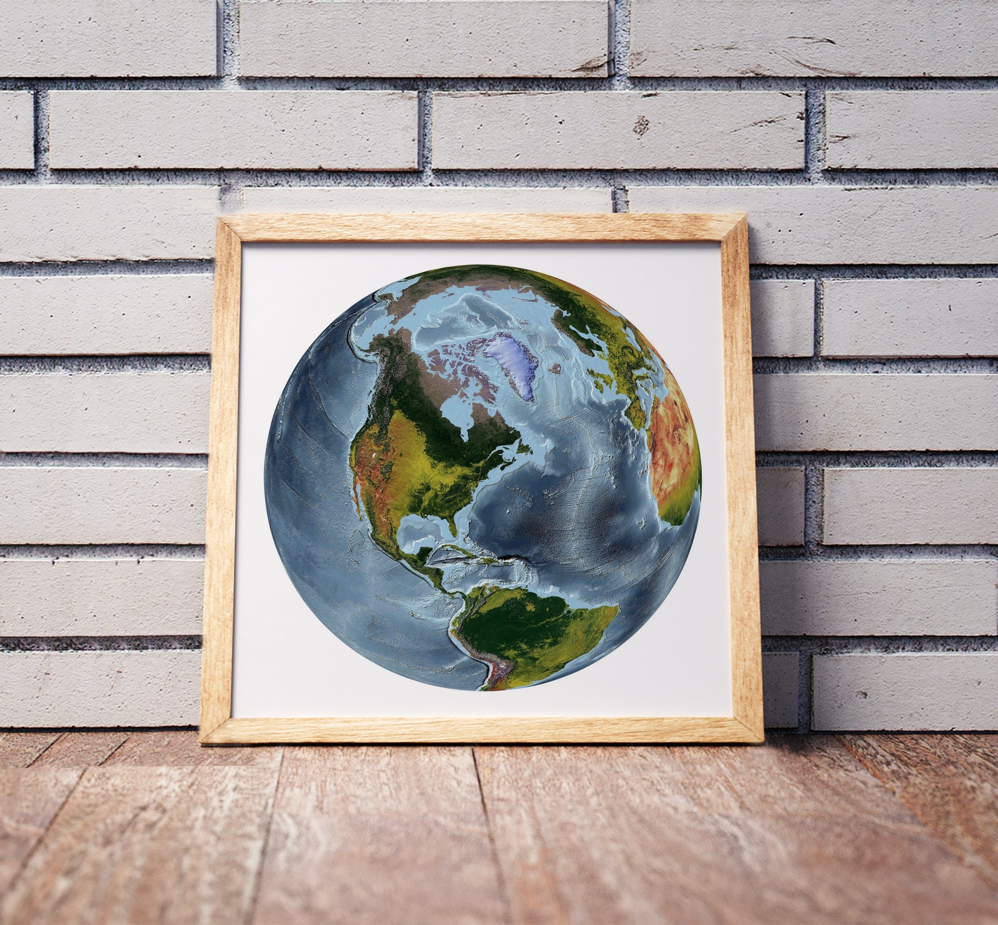 The World From Space projection shaded relief world map in a wooden frame. Realistic terrain art print for home decor, displayed against a brick wall