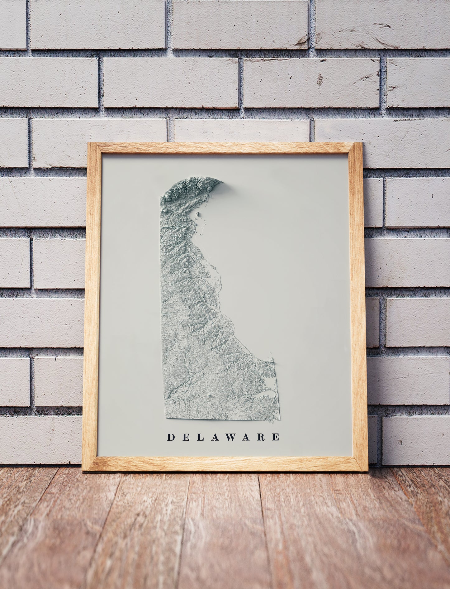 Delaware shaded relief map in a wooden frame. Realistic terrain art print for home decor, displayed against a brick wall