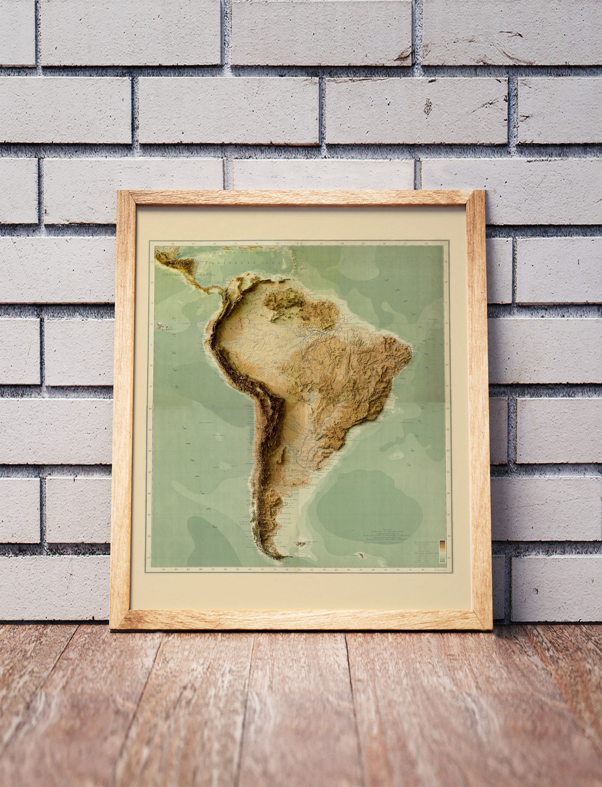 Vintage South America shaded relief map in a wooden frame. Realistic terrain art print for home decor, displayed against a brick wall